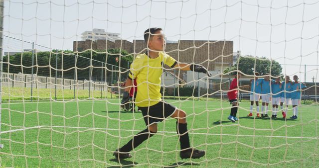 Young boy wearing goalkeeper gear standing ready to defend goal during a soccer match. In the background, teammates and opponents standing on the field. Perfect for depicting children's sports activities, teamwork, and outdoor competition.