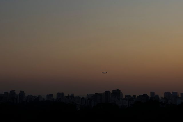 Silhouetted airplane flying against a backdrop of a city skyline lit by the glow of sunset. Ideal for travel promotions, city living articles, or illustrating the calmness of twilight in urban settings.