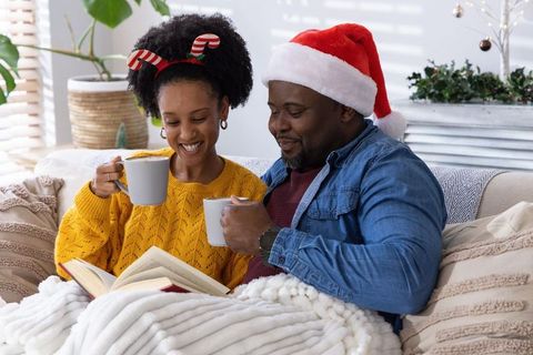 African american couple reading and sharing hot cocoa on cozy sofa wearing santa hat