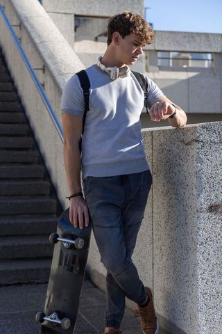 Young skateboarder leaning on concrete railing checking watch with headphones in sunlight