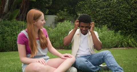 Young diverse couple relaxing on lawn sharing headphones and enjoying music outdoors