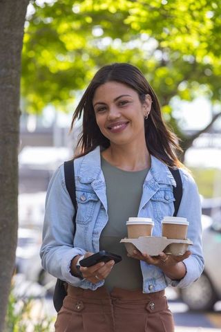 African American woman smiling holding two to-go coffees and smartphone on sunny street