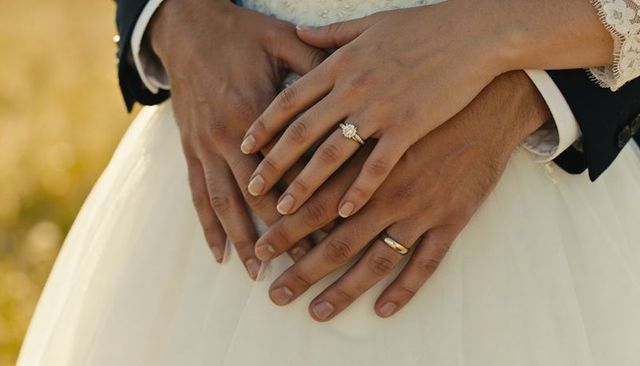 Couple holding hands showing halo engagement ring and gold wedding band over white gown in meadow