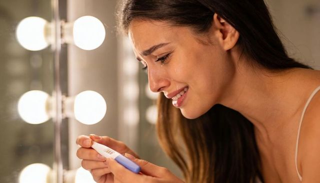 Tearful young woman checking pregnancy test at lit vanity mirror, reacting in tears