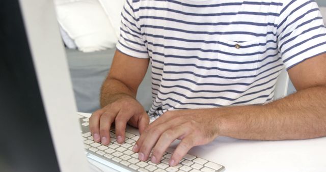 A man is typing on a keyboard while sitting at a desk. He is wearing a casual striped shirt. This image can be used for representing work, remote job, casual work environment, technology use, or home office setup.