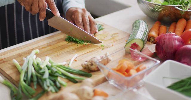 Person chopping fresh vegetables on wooden cutting board in modern kitchen. Green onions, carrots, red bell pepper and ginger are being prepared next to professional knife, indicating healthy eating and skillful cooking. Ideal for use in content about healthy meal preparation, professional cooking techniques, or culinary classes.