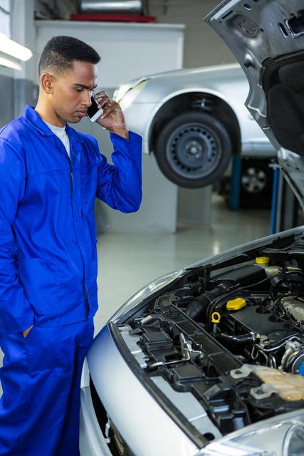 Mechanic in blue overalls talking on a mobile phone while inspecting a car engine in an auto repair garage. Ideal for use in articles or advertisements related to automotive services, car maintenance, professional repair services, and communication in the workplace.