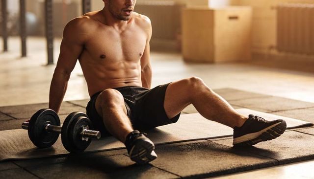 Shirtless male athlete resting on mat after intense dumbbell workout in sunlit gym
