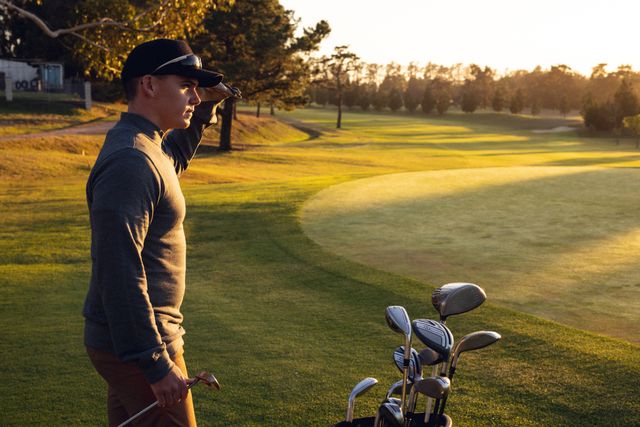 Side view of caucasian young man with golf clubs looking away while standing on field golf course. Sunset, golf, unaltered, nature, sport and weekend activities concept.