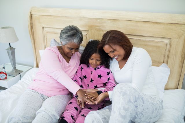 Three generations of women, including a grandmother, mother, and daughter, bonding and smiling while sitting on a bed in a cozy bedroom. They are forming a hand stack, symbolizing unity and love. This image is perfect for use in family-oriented advertisements, lifestyle blogs, and articles about family relationships and bonding.