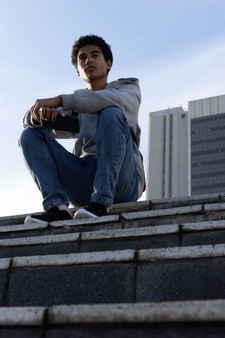 Teenager sitting on concrete steps holding smartphone, gazing at urban skyline