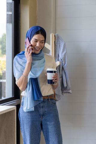 Sunlit woman wearing blue hijab talking on smartphone while holding coffee cup