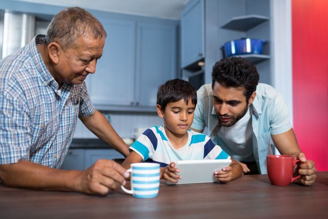 Family members gathered around a digital tablet in a kitchen, engaging in a shared activity. Grandfather, father, and son are bonding and learning together. Ideal for illustrating family togetherness, technology use in everyday life, and multigenerational interactions.