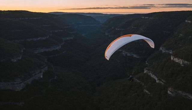 Paraglider soaring over deep forested canyon at sunset with white wing and orange trim