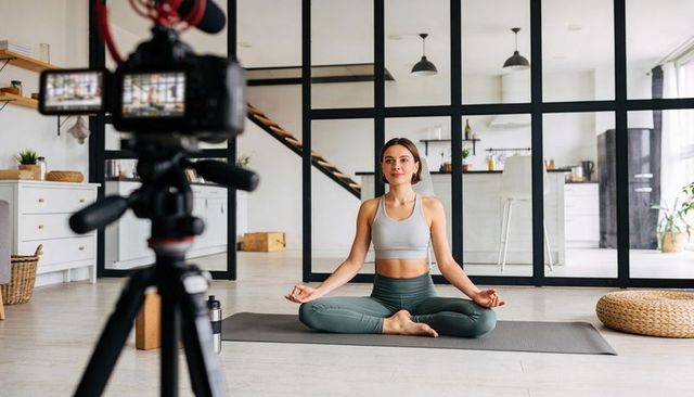 Filming yoga tutorial: woman meditating on mat in sunlit loft wearing workout clothes
