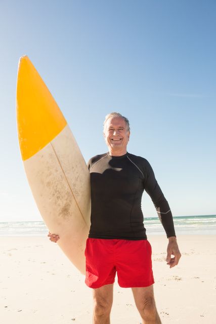 Senior man standing on sandy beach holding surfboard, smiling under sunny sky. Ideal for promoting active lifestyle, retirement activities, beach vacations, and healthy living. Perfect for travel brochures, fitness campaigns, and summer holiday advertisements.