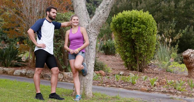 A young couple is seen relaxing and chatting near a tree after completing an outdoor workout in a park. Both are dressed in sportswear, suggesting an active lifestyle focused on fitness and healthy living. This image can be used for promoting outdoor exercise routines, active lifestyles, fitness apps, or advocacy for spending time in nature.
