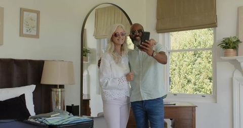 Mature couple taking selfie in bright bedroom with mirror sunglasses and warm decor