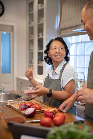 Senior asian couple preparing vegetables and sharing wine in cozy daylight kitchen