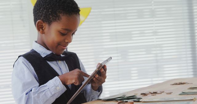 Young boy wearing a bow tie engaged with a tablet, surrounded by coins and bills on a table in front of him. Ideal for themes related to financial literacy for kids, early education on money management, technology in learning, or future financial planning content.