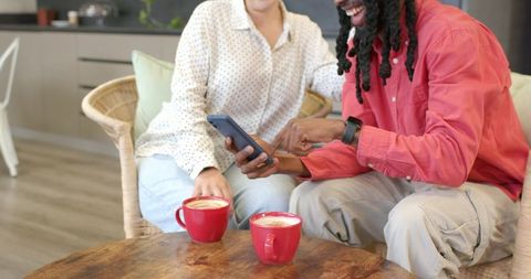 Diverse friends sharing coffee and scrolling smartphone in cozy modern lounge, casual conversation
