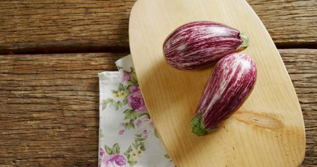 Two striped, purple eggplants placed on a light wooden board with a floral-patterned napkin underneath. Background features rustic wooden surface. Great for food blogs, cooking magazines, and culinary websites focused on fresh produce and recipes.