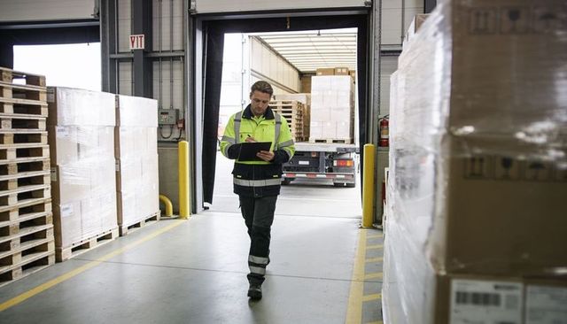 Warehouse worker checking clipboard while walking through loading dock between pallets and trailer