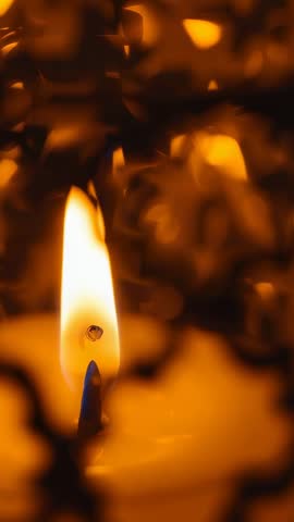 Vertical close-up candle flame leaning in breeze with melting wax droplet and warm bokeh