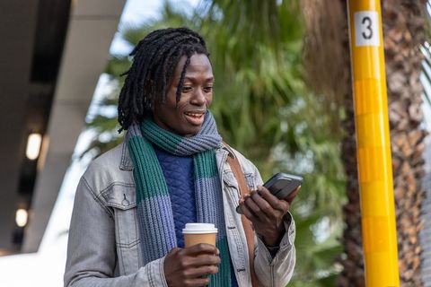 African american commuter checking smartphone holding coffee by transit pole with palm trees