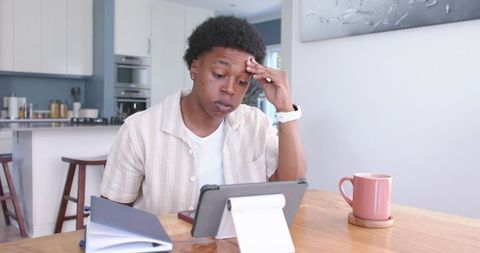 Young man studying on tablet at kitchen table looking stressed, remote learning focus home