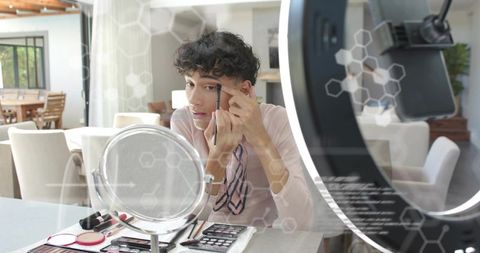Curly-haired man applying eyebrow makeup at vanity with ring light and streaming setup