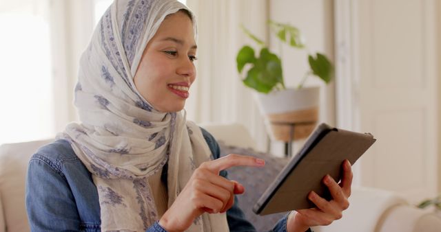 Middle Eastern woman wearing hijab using tablet for communication, sitting comfortably at home. Ideal for use in technology, communication, lifestyle, and cultural representation topics.