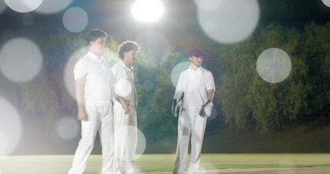 Three cricketers standing on night pitch under floodlights wearing white uniforms