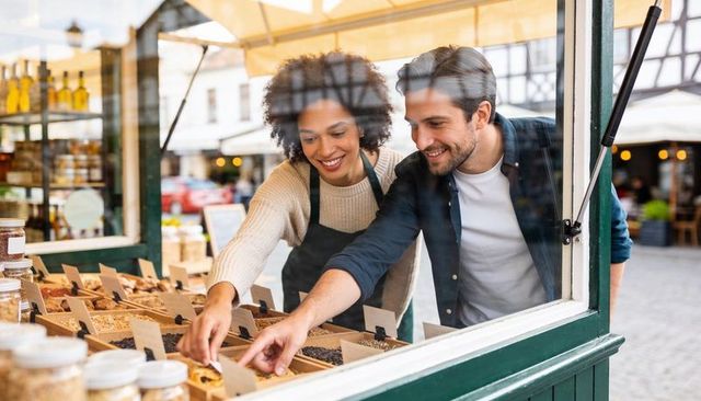 Couple smiling while selecting bulk goods at outdoor market stall with jars and canopy