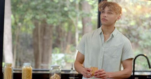 Young man holding wooden cup, standing in sunlit kitchen