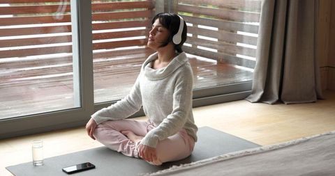 Woman meditating with headphones near balcony in cozy room