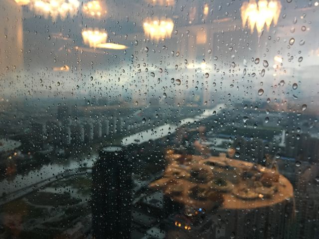 Rain covered window overlooking urban cityscape with skyline in background. Indoor lights reflecting on glass add warmth to the scene. Ideal for use in articles about rainy weather, urban life, or reflective moments in city environments.