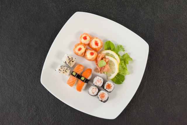 This image features an assorted sushi platter served on a white plate against a black background. It includes various types of sushi such as nigiri, rolls, and sashimi, accompanied by wasabi, ginger, lemon, and lettuce. Ideal for use in food blogs, restaurant menus, culinary websites, or advertisements promoting Japanese cuisine.