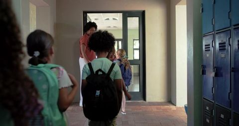 African american teacher greeting elementary students walking into corridor with backpacks