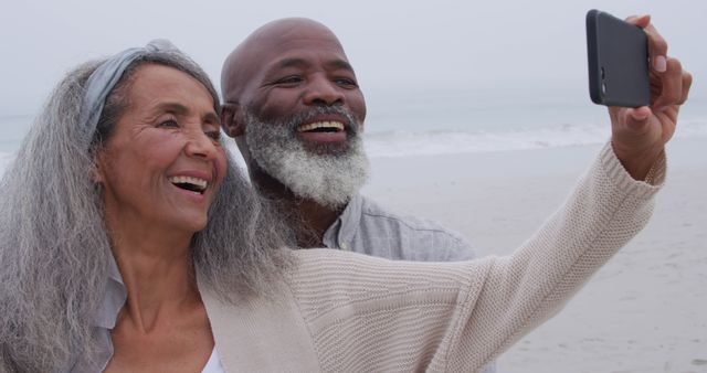 Elderly couple enjoys capturing a moment together on the beach by taking a selfie. Ideal for content related to senior activities, travel, lifestyle, and family moments.