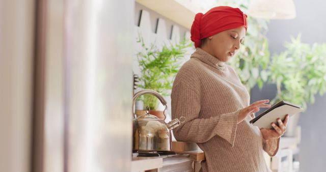 Image of smiling biracial woman in hijab using tablet standing in kitchen at home. Happiness, communication, relaxation, inclusivity and domestic life.