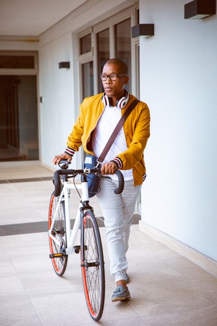Young biracial male business creative arriving at workplace with bike. Wearing casual yet stylish clothing, including headphones and a messenger bag. Ideal for concepts related to urban commuting, modern work environments, independent professionals, and sustainable transportation.