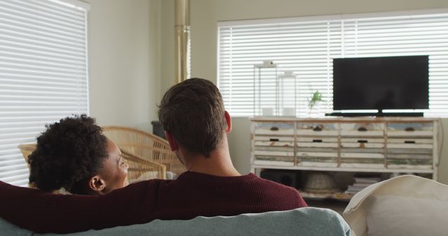 A couple seen from behind relaxing on couch, watching television in cozy living room. Background includes modern furniture and white blinds covering windows. Suitable for themes on relationships, leisure time, home life, and modern living setups.