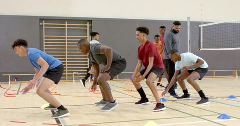 Diverse male athletes doing agility drills on indoor court with coach observing teamwork