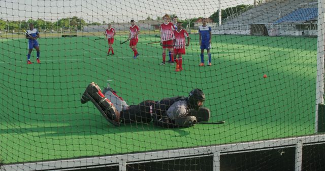 Rear view of a Caucasian male field hockey goalkeeper, diving to stop the ball with his hockey stick during the game on the hockey pitch, with his teammates and opponents in the background