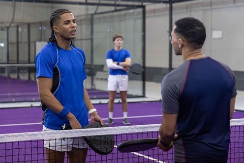 Diverse male athletes talking across net on indoor padel court with purple playing surface