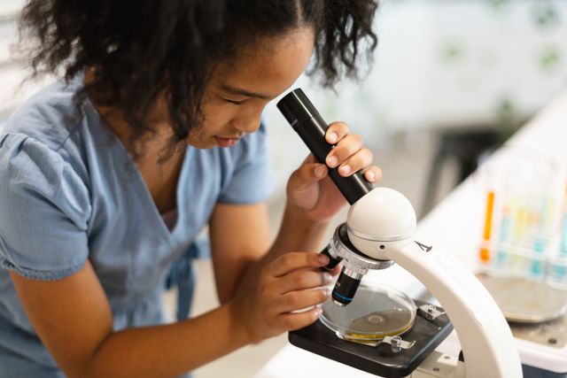 Biracial elementary schoolgirl analyzing chemical under microscope during chemistry practical. unaltered, education, laboratory, stem, scientific experiment and school concept.