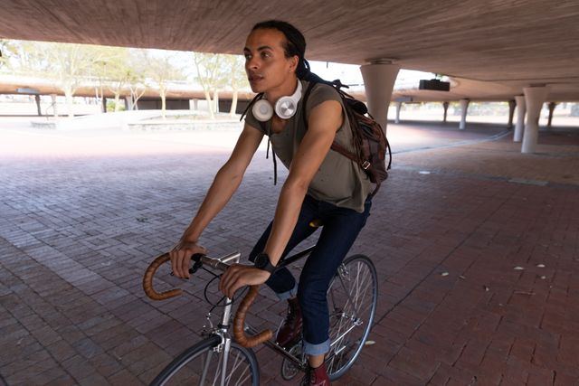 Biracial alternative man with dreadlocks and headphones out and about in the city on a sunny day, riding bike under bridge. Urban trendy man on the go.