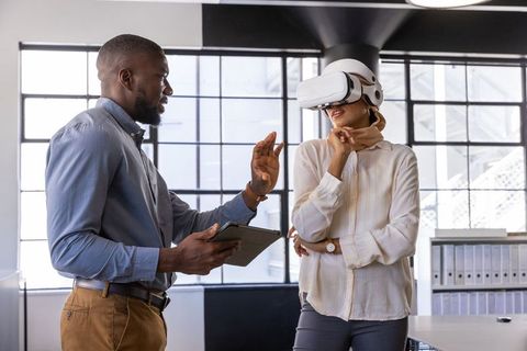 Two professionally dressed coworkers in an office, one using a VR headset while the other discusses insights from a tablet. This image showcases modern business environments embracing cutting-edge technology. Ideal for illustrating technology integration, modern teamwork dynamics, digital transformation, and collaborations in business settings.