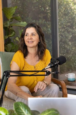 Mature woman hosting cozy home podcast wearing mustard blouse surrounded by plants